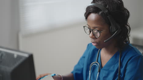 A-black-woman-doctor-wearing-headphones-sits-at-a-table-with-a-computer-and-takes-calls-from-patients-looks-at-their-medical-records-and-enters-them-into-the-clinic-schedule
